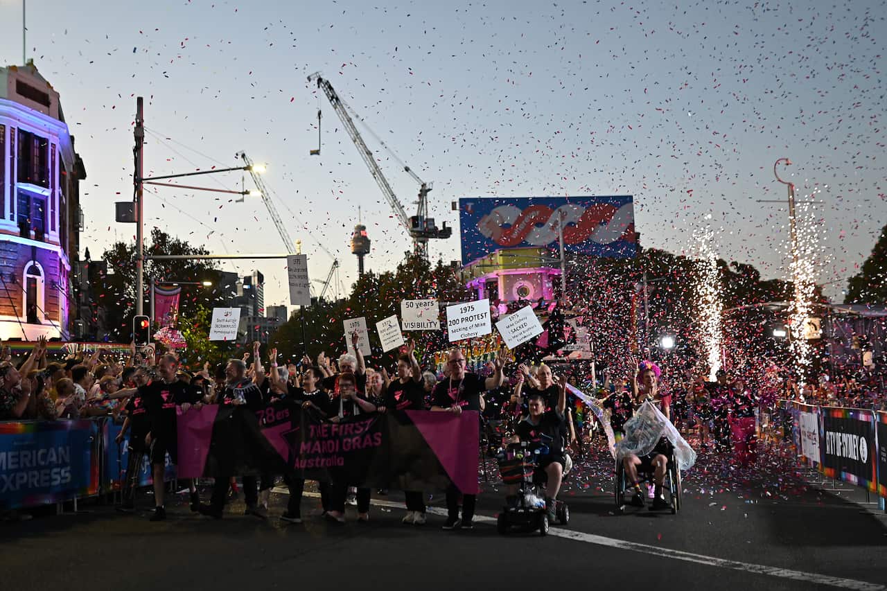 A crowd of people walk or roll in wheelchairs/mobility scooters holding a black and pink banner that reads "78ers - the first Mardi Gras in Australia". There is much confetti being thrown in the air.