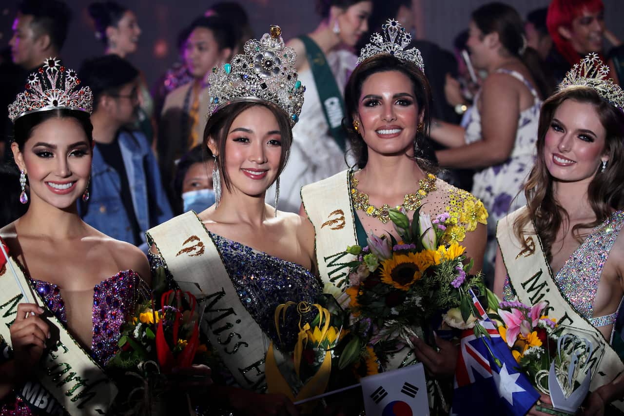 Four women wearing makeup, sashes and gowns smile. They are all holding flowers.