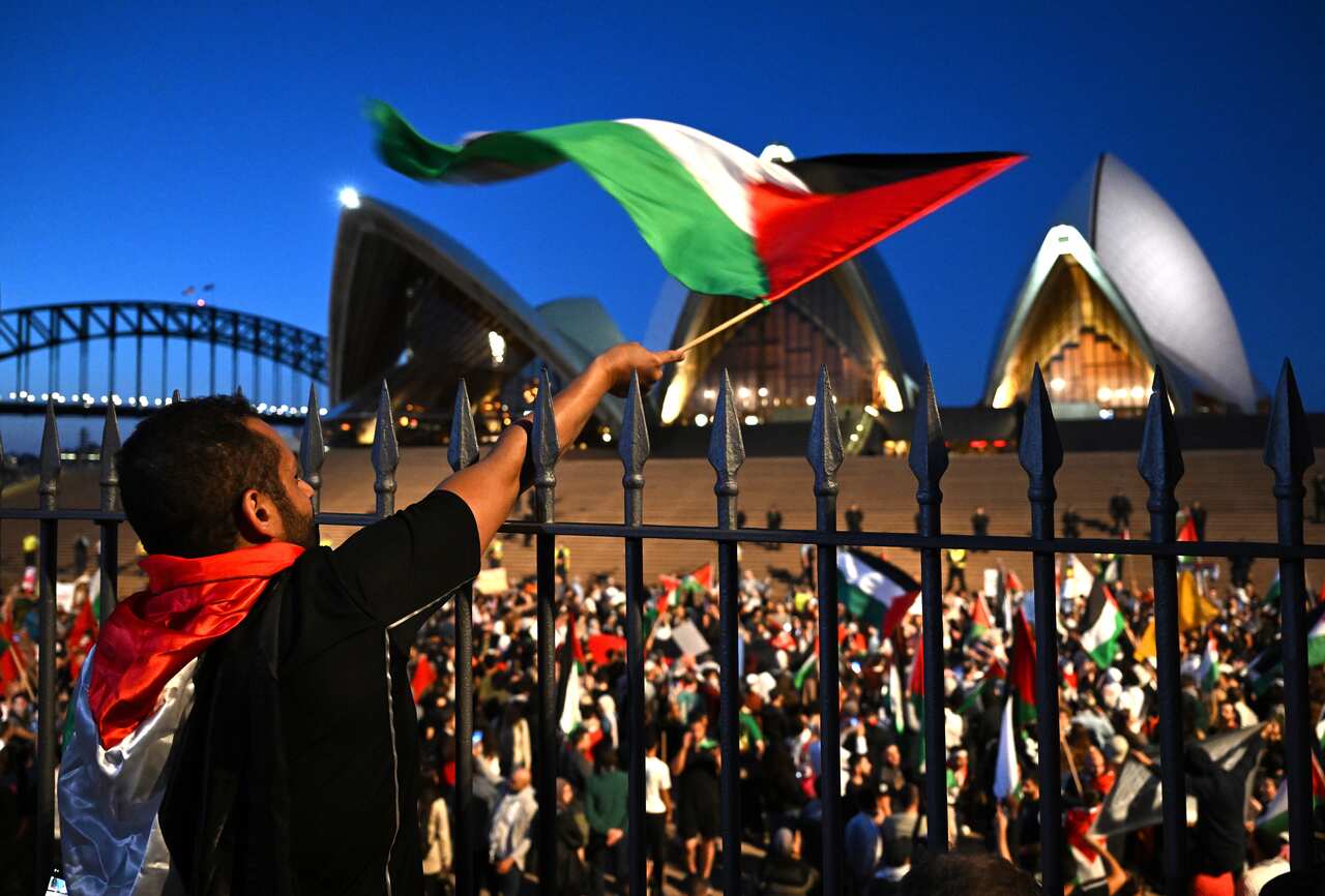 A man leaning on a fence waves a Palestinian flag. A crowd below is in front of the Sydney Opera house.