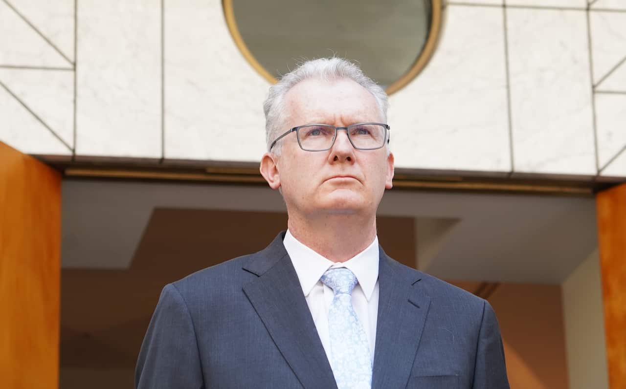 A man in a dark blue blazer, standing in front of a white building with brown doors.