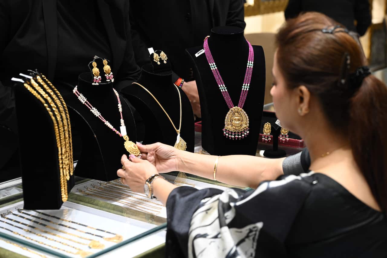 A woman looking at a gold necklace on a black display bust at a jeweller's. The bust is one of four featuring gold necklaces and placed on a display cabinet also featuring gold necklaces.