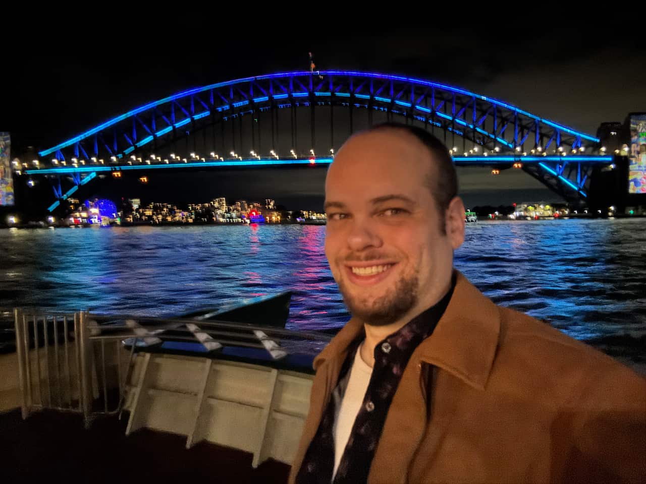 A man smiling in a selfie in front of the Sydney Harbour Bridge at night