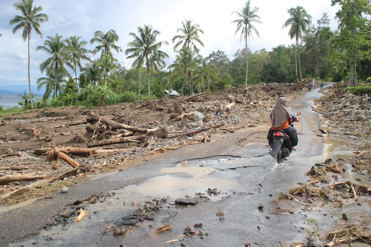 People drive on a motorbike on a road that's partly been washed away by flooding. Trees and other debris are scattered either side. 