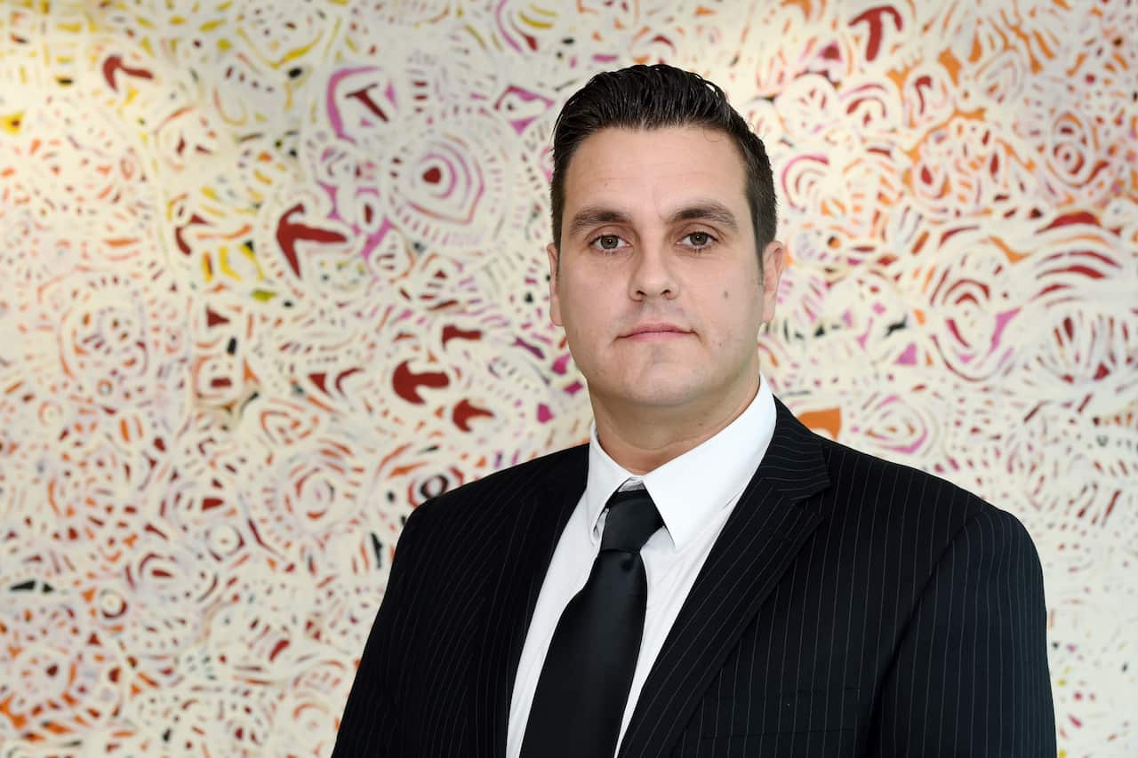 A man in a suit stands in front of a colourful background with a neutral expression on his face.