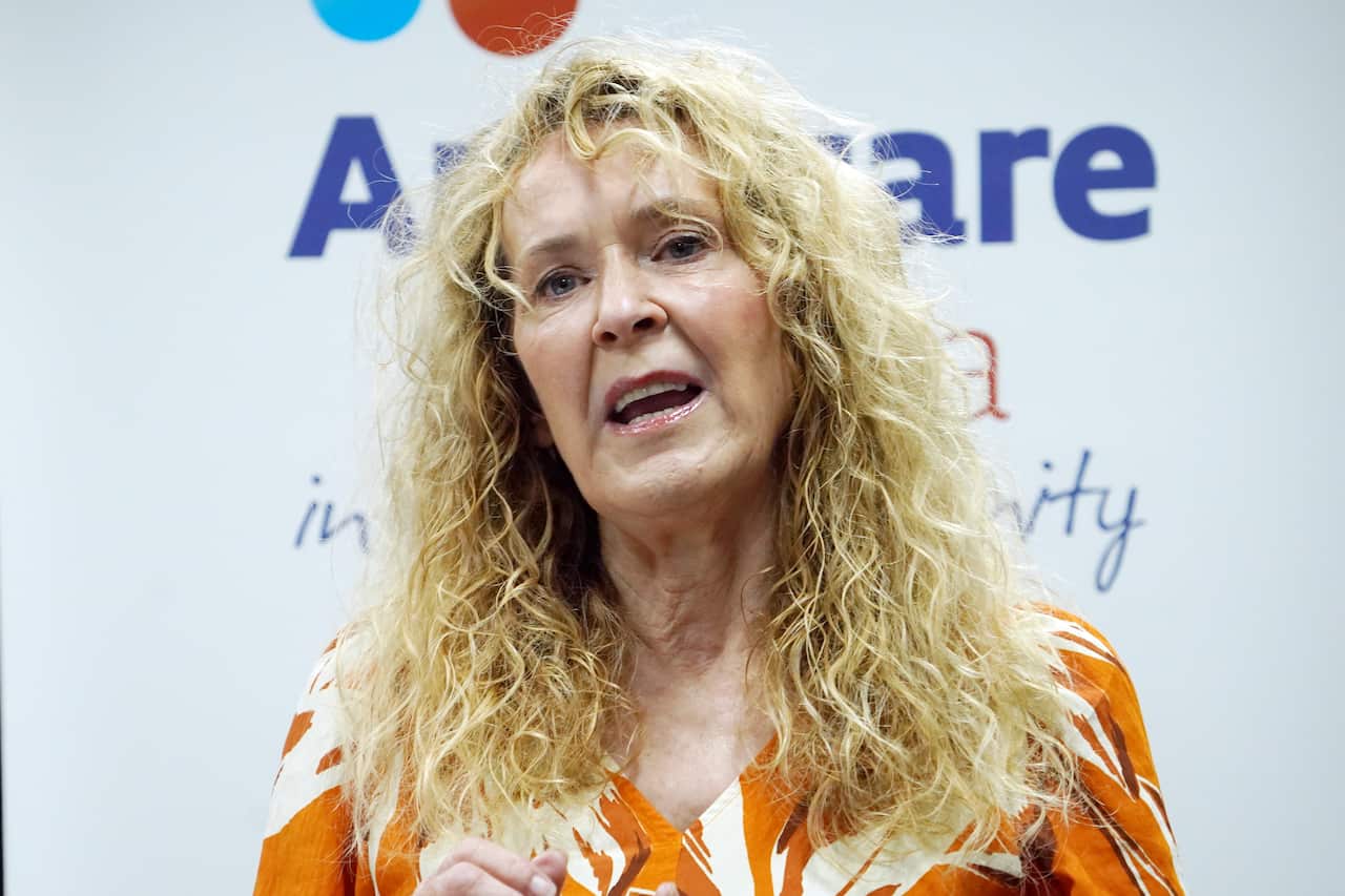 A woman with long, blonde curly hair speaks. She is standing in front of a white media wall with Anglciare's logo on it