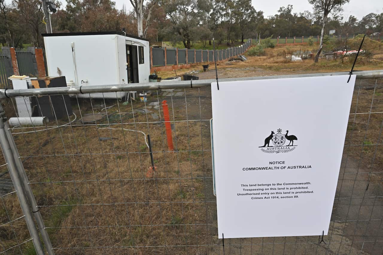 An empty block of land with a small white demountable building on it. A temporary chain link fence with a sign saying the land belongs to the Commonwealth of Australia.