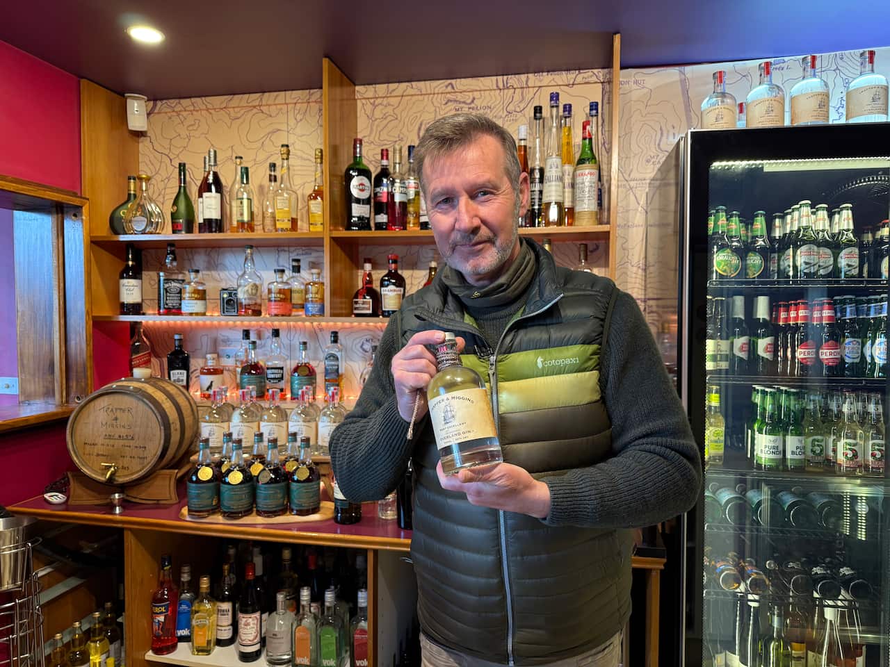 A middle aged white man holding up a bottle of gin. Behind him is a bar with various alcoholic drinks. 