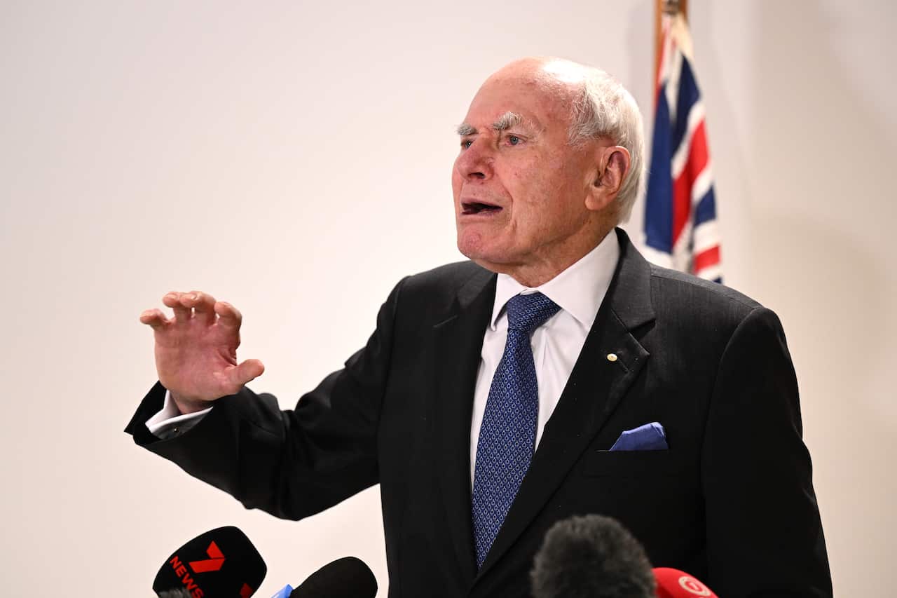 John Howard in a suit and tie speaking from behind microphones