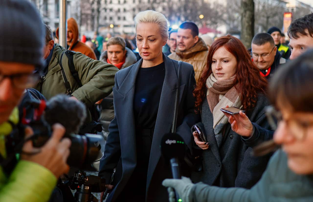 A woman with short grey hair, in a blue coat, flanked by reporters as she walks outside.