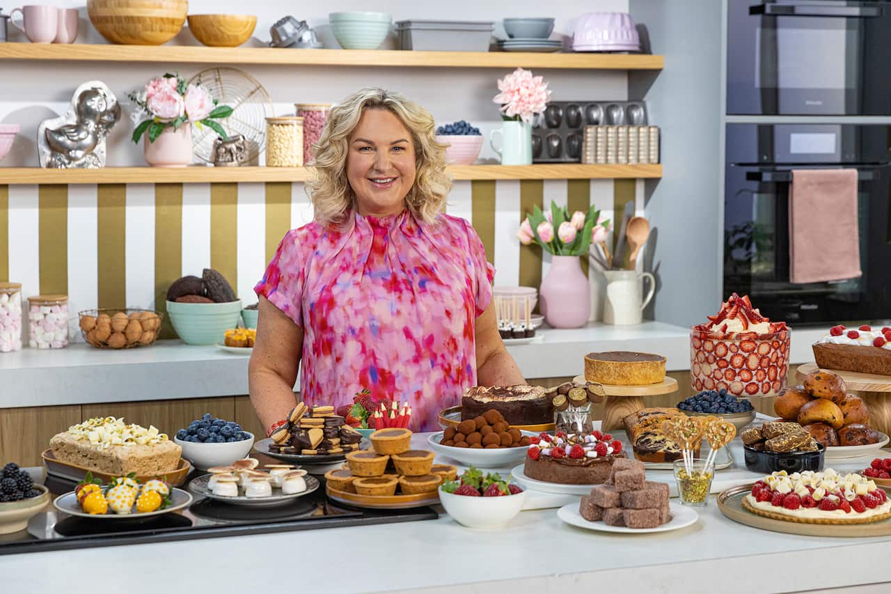 A smiling woman in a patterned pink dress stands in a kitchen, behind a bench which displays many different baked and cooked chocolate dishes, including cakes, tarts, biscuits and buns.
