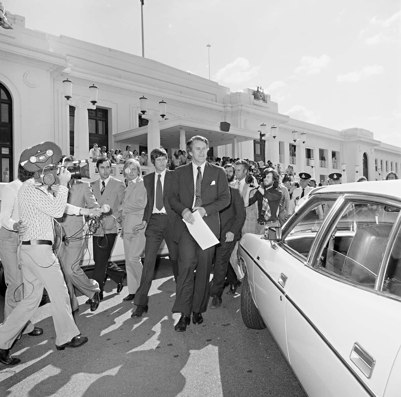 A man in a suit holding a document walks away from Old Parliament House towards a parked car, with reporters and cameras following him.