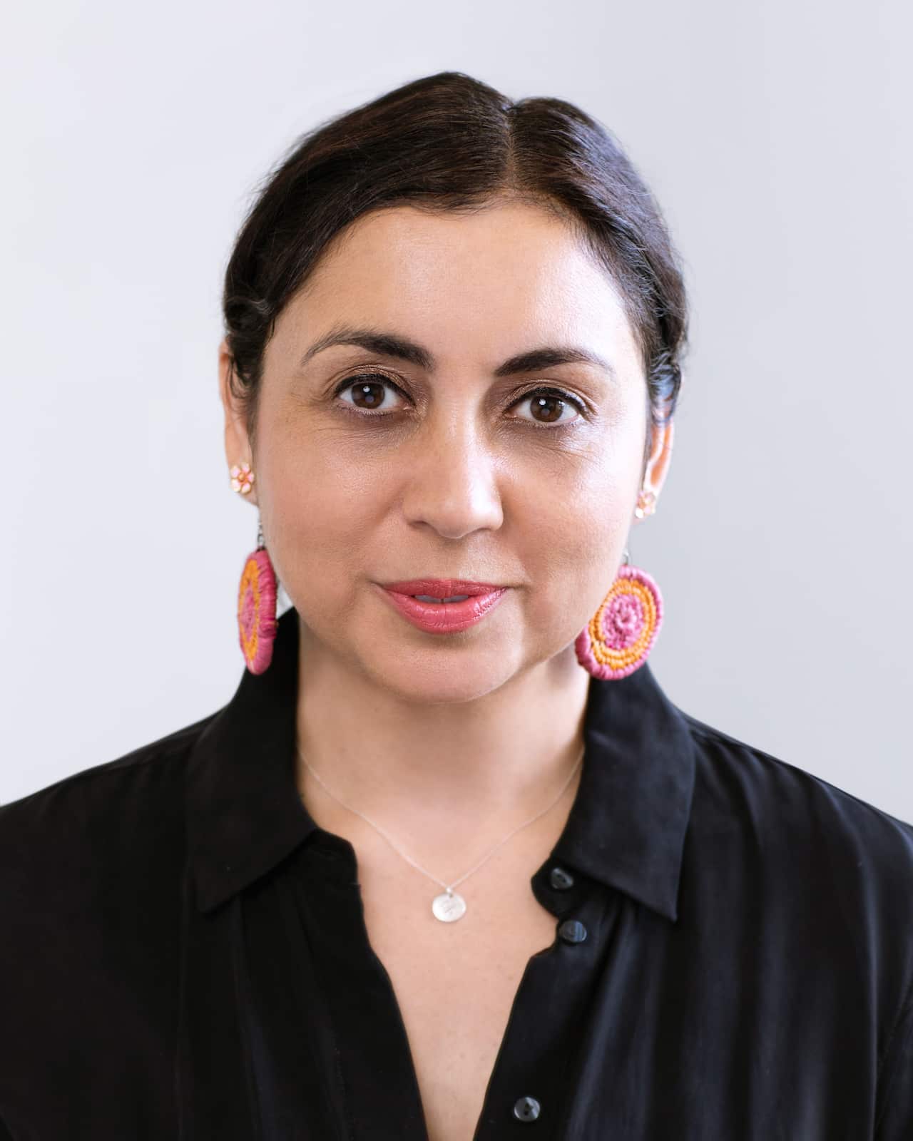 A woman wearing a black shirt and colourful earrings against a white background.