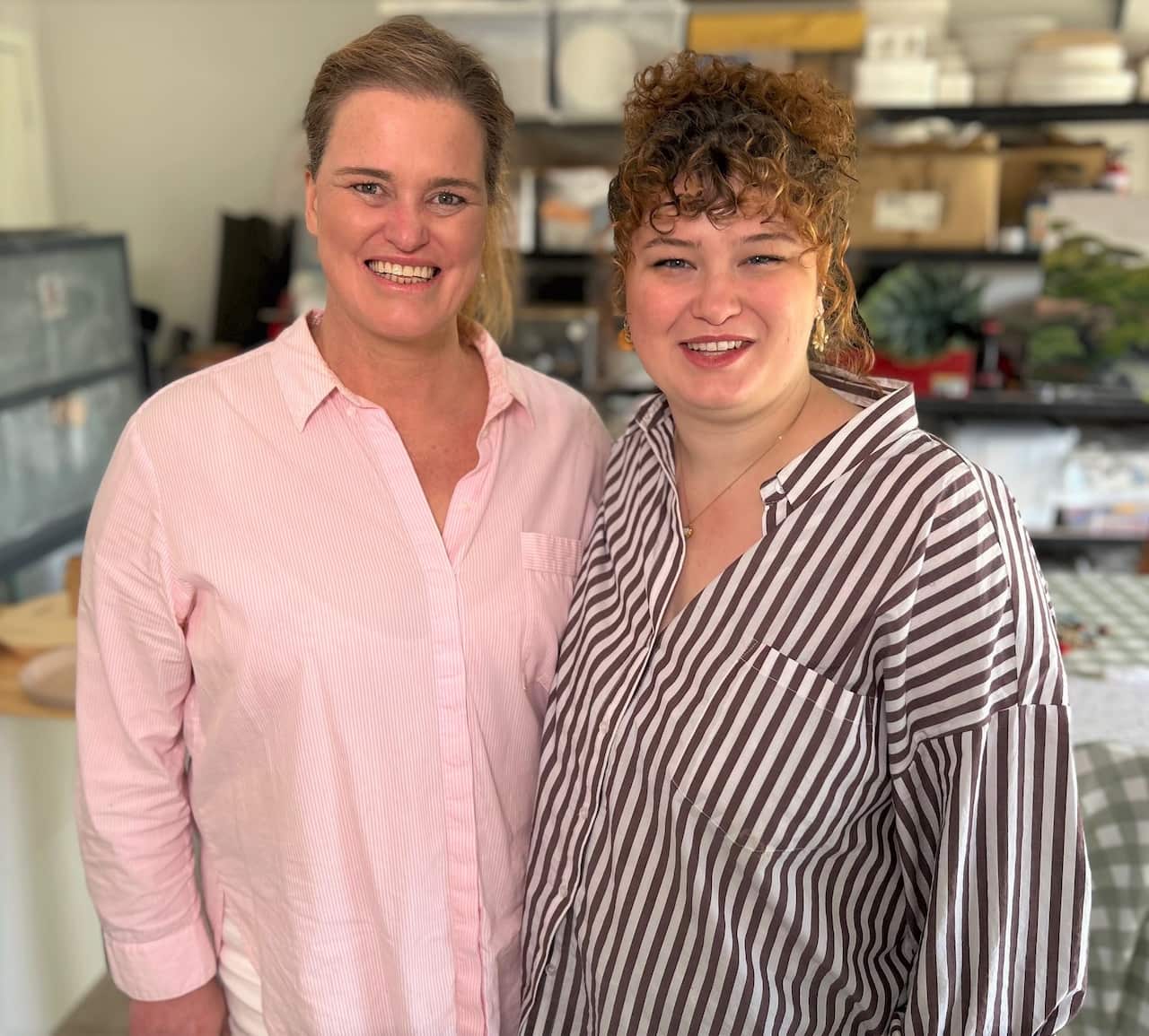 A woman with brown hair wearing a pink shirt stands next to a younger woman wearing a striped shirt.