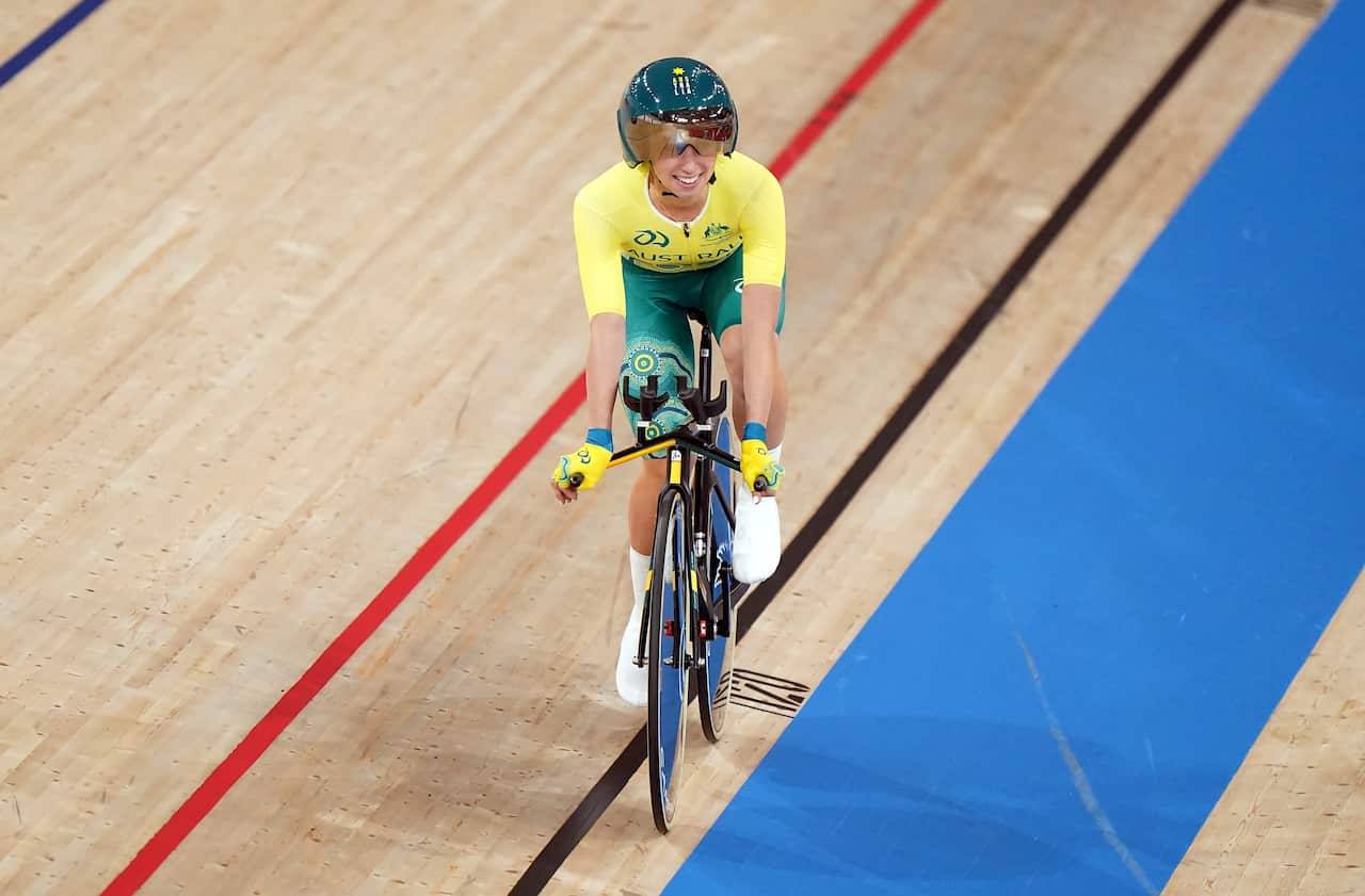 A woman wearing a yellow top, green cycling shorts and a green helmet riding her bike on a wooden indoor cycling track