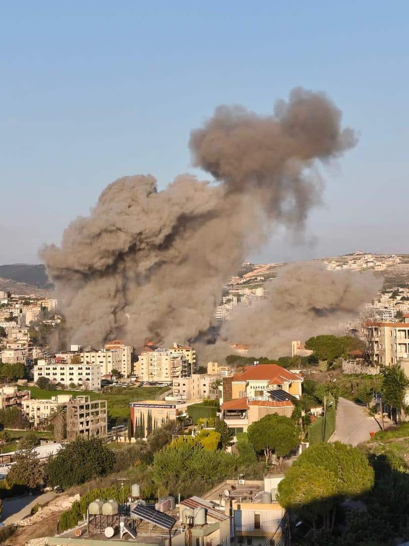 Large plumes of grey and brown smoke rise from explosions amid a densely populated residential area with multi-storey buildings and rolling hills in the background.