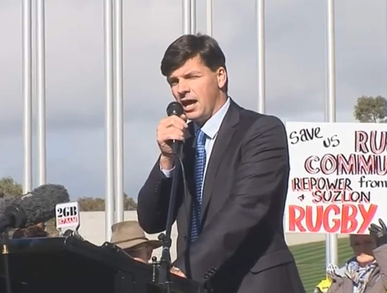 Angus Taylor fala ao microfone em um pódio ao ar livre com sinais de protesto visíveis ao fundo.