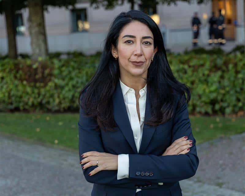A dark haired woman in a suit stands with her arms crossed, a slight smile on her face. 