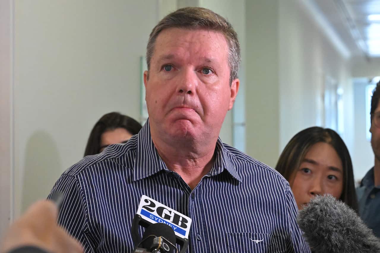 A man in a blue striped shirt speaking in front of microphones
