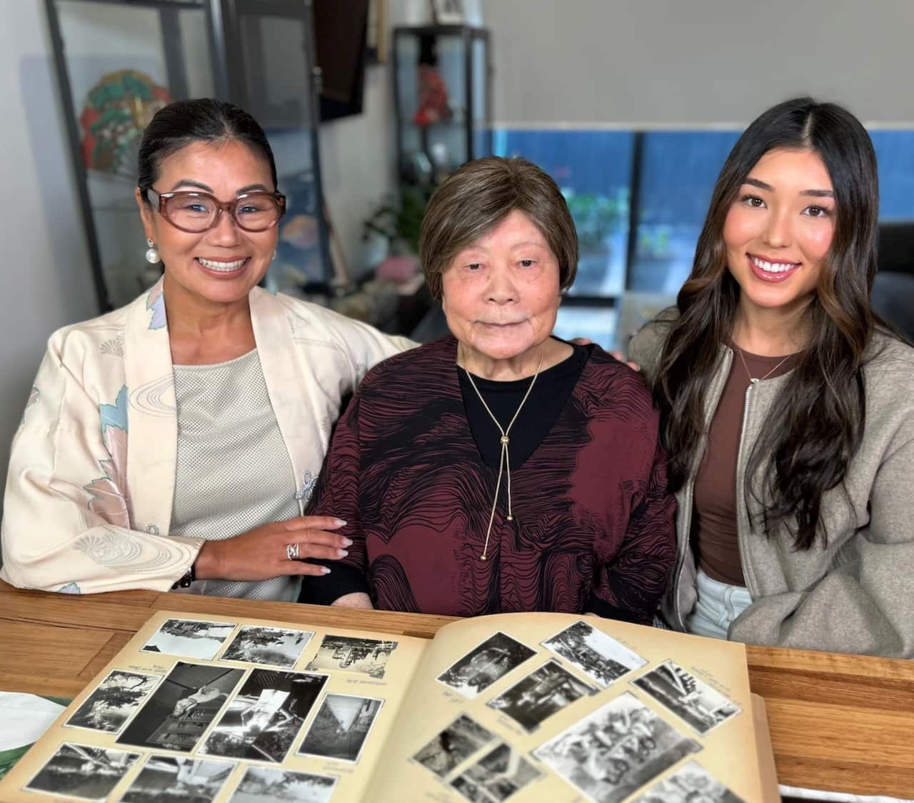 An elderly woman, flanked by two young women on either side, smiles as she sits at a wooden table with a photo album placed on it.