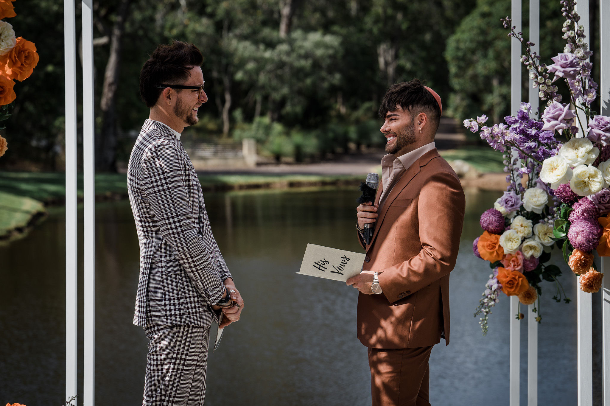 A gay couple stand facing each other on their wedding. The man on the left is in a black and white checkered suit and smiling. The man on the right is wearing a brown suit and kippah, holding a microphone and a booklet with text reading 'his vows'. 