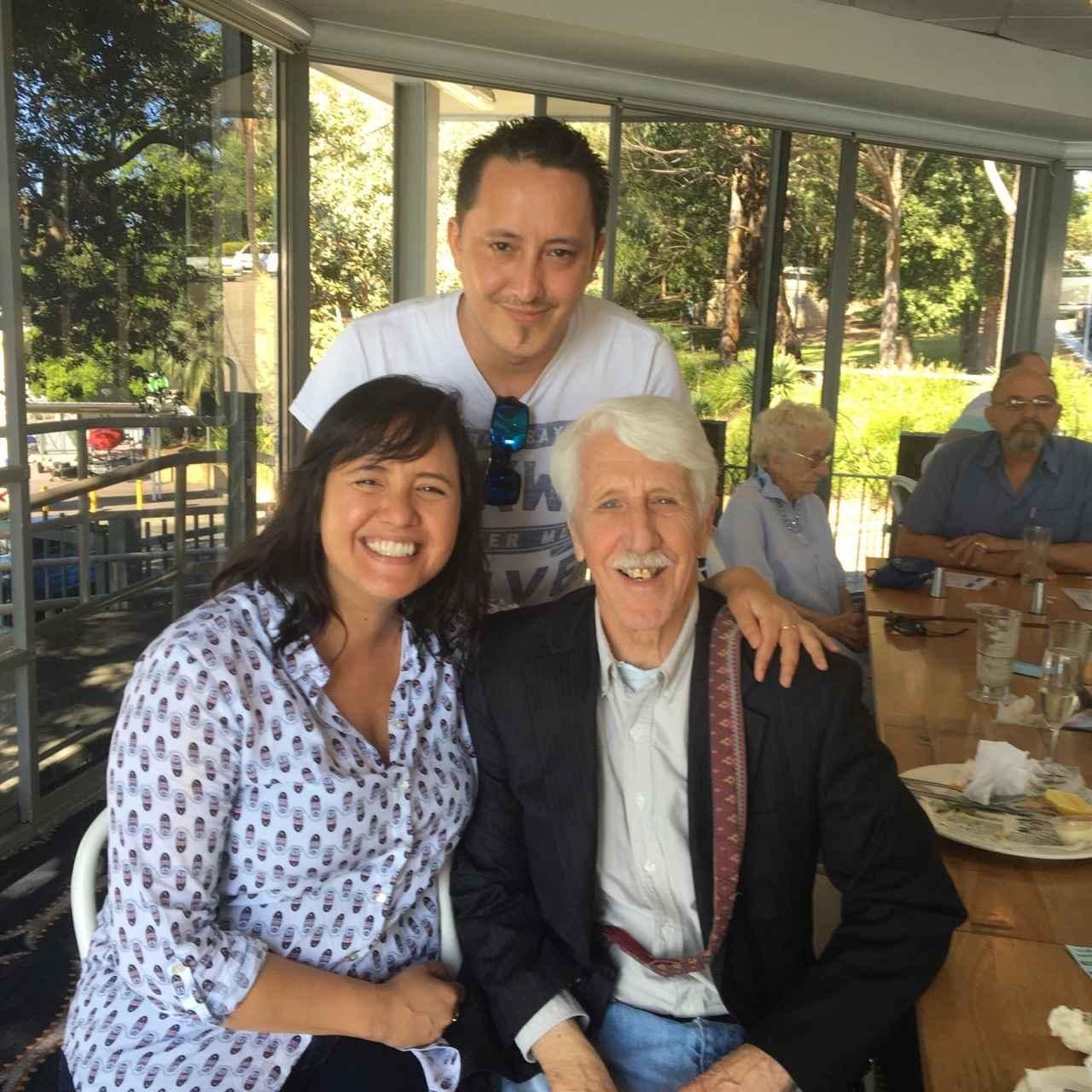 Um homem parado atrás de um homem mais velho e de uma mulher mais jovem, todos sorrindo.