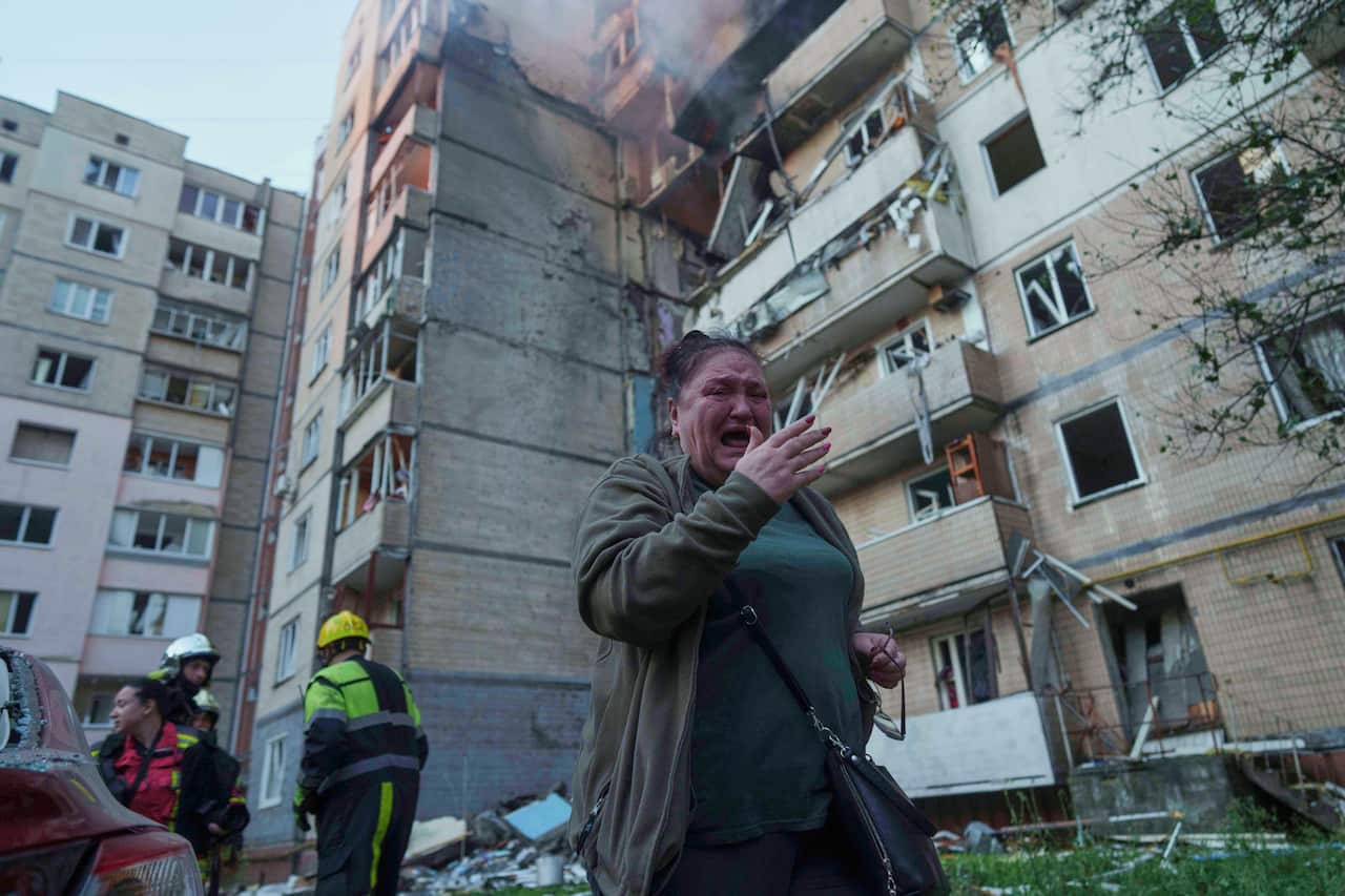 A woman cries as she flees a damaged building. There are emergency personnel standing behind her.