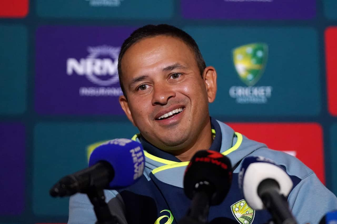 Usman Khawaja sits smiling behind several microphones at a press conference, wearing an Australian cricket team hoodie against a backdrop featuring Cricket Australia and NRMA Insurance logos.