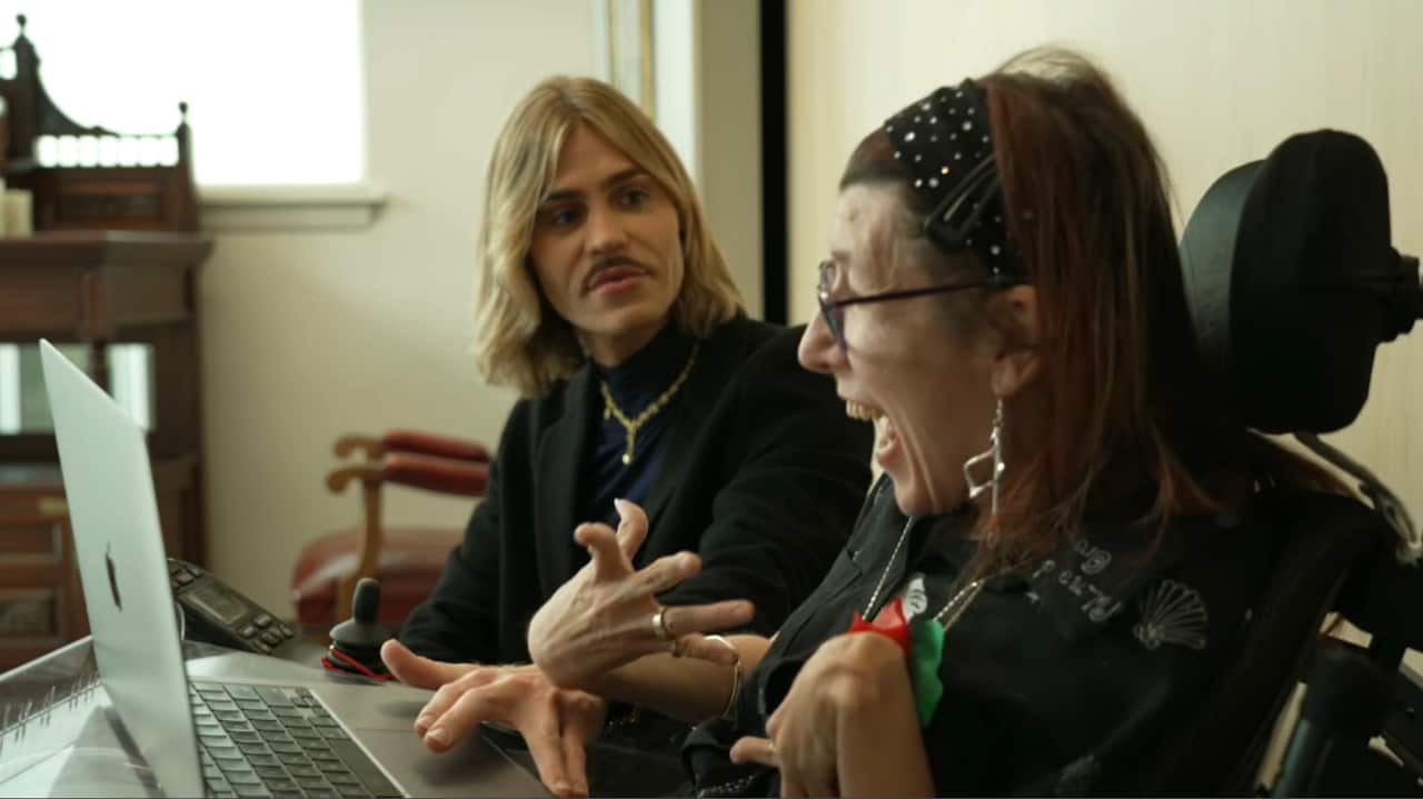 A person with blonde hair and a black top using a laptop alongside a person with red hair sitting in a wheelchair.