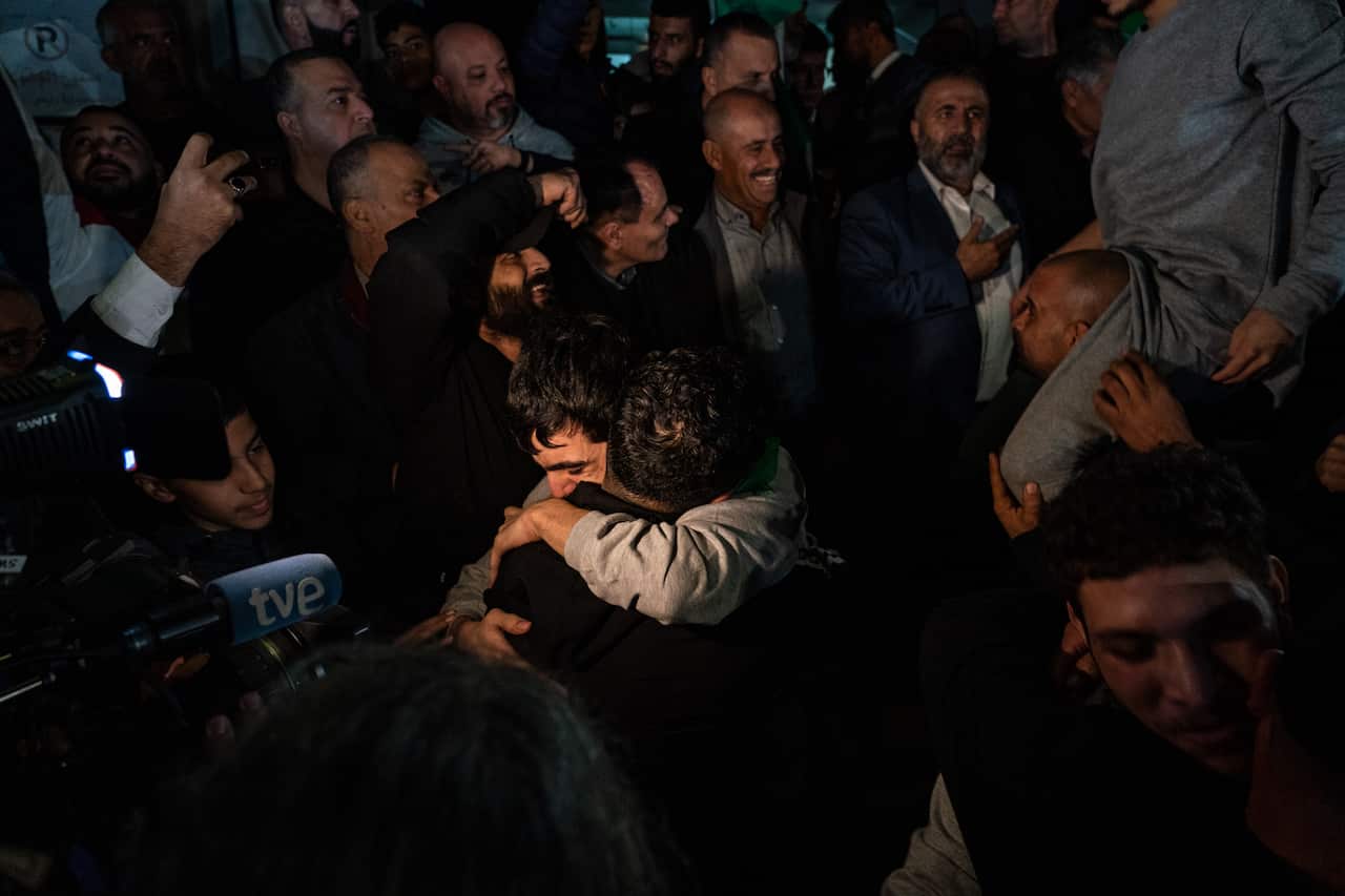 Two men embrace tightly at the centre of a dense, emotional crowd filled with onlookers and news cameras at night.