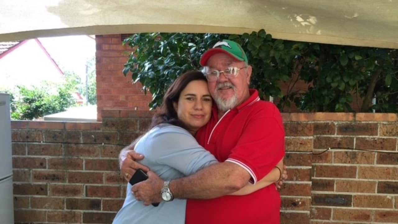 a young woman with long brown hair hugging an older man wearing a green cap with a white rabbit symbol and a red t-shirt standing in front of a brick fence