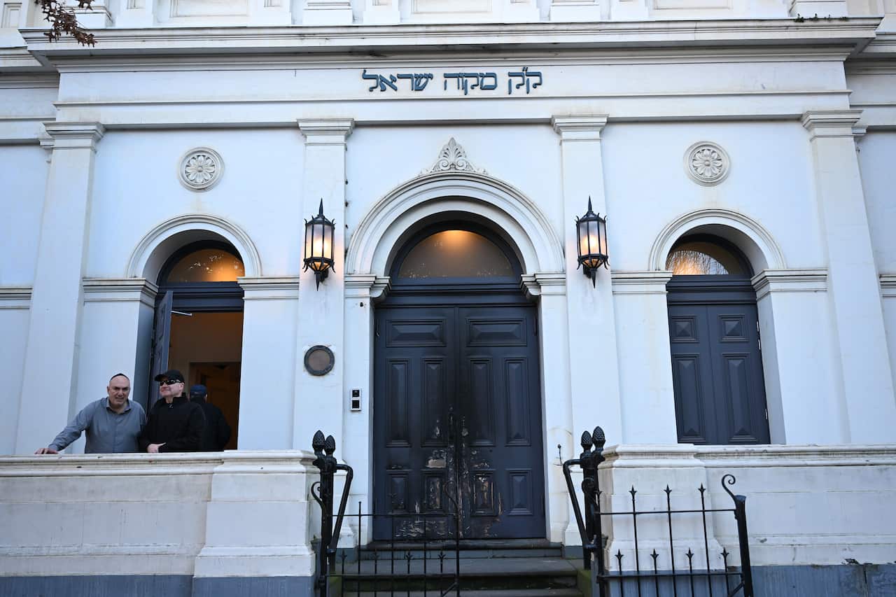 Two men stand outside a synagogue with dark doors and Hebrew writing above the entrance.