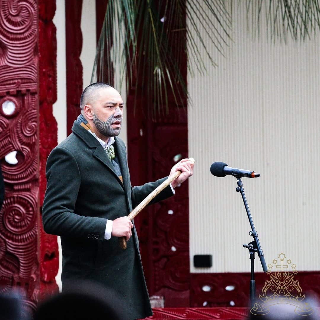 A man wearing a grey coat, speaking at a microphone and holding a wooden stick. He has a traditional facial tattoo on the lower half of his face.