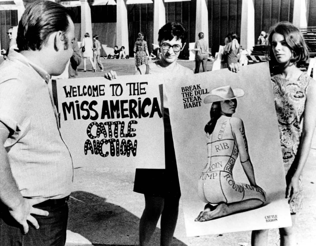 A black and white picture of female protesters holding two signs. One says 'Welcome to the Miss America cattle auction' and the other has a picture of a naked woman wearing a cowboy hat. Someone has drawn on her body, dividing it into sections like a steak, with text reading 'rib, loin, rump, chuck'. Above her is text reading 'Break the dull steak habit'