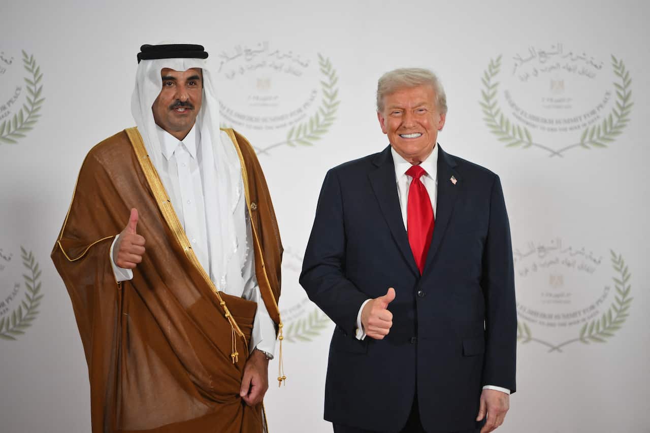 A man in a traditional Arab robe and another in a black suit and a red tie give thumbs up as they smile for the camera.