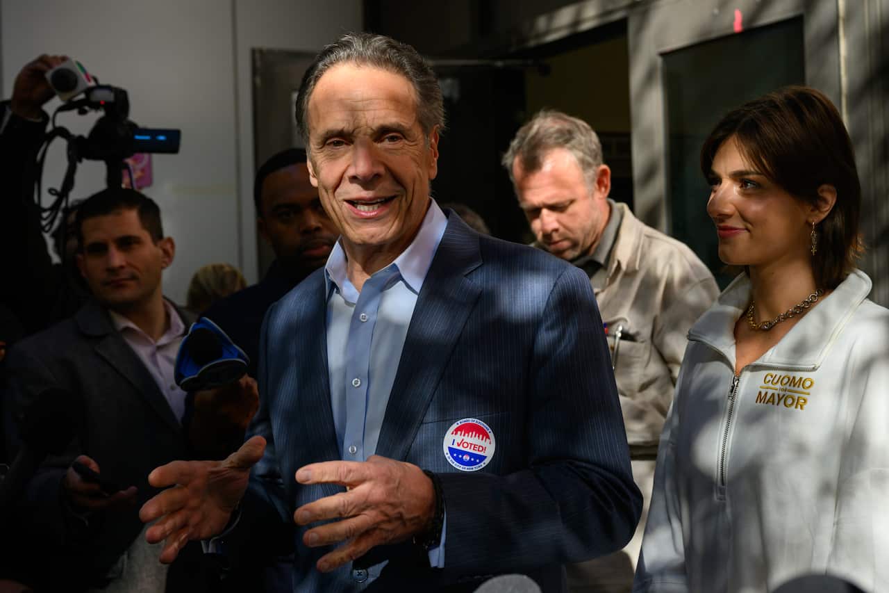 A middle-aged white man wearing a blue suit jacket and shirt. He's speaking and holding has hands half-raised and in front of him. A group of people, including press, surround him.