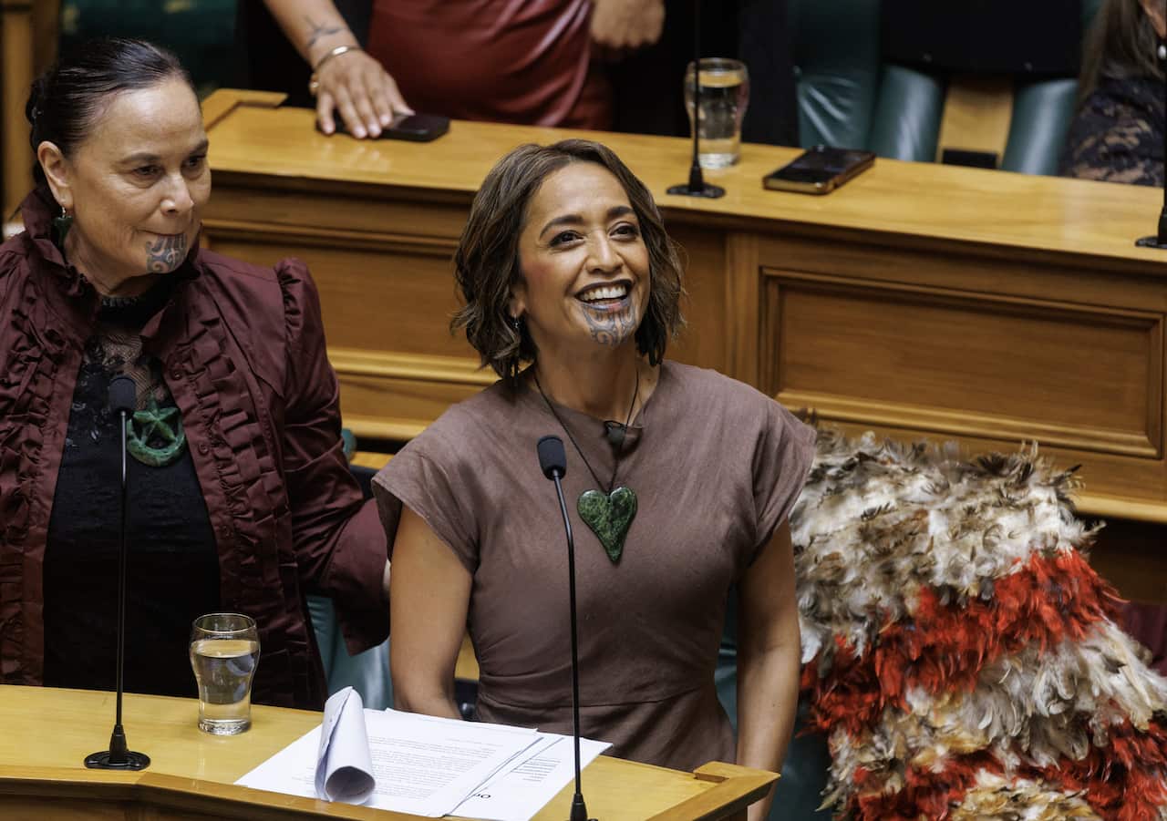 A smiling woman with a traditional tattoo on her chin, sitting inside. She has a feathered cloak beside her, and a colleague, also with a facial tattoo, stands to her side.