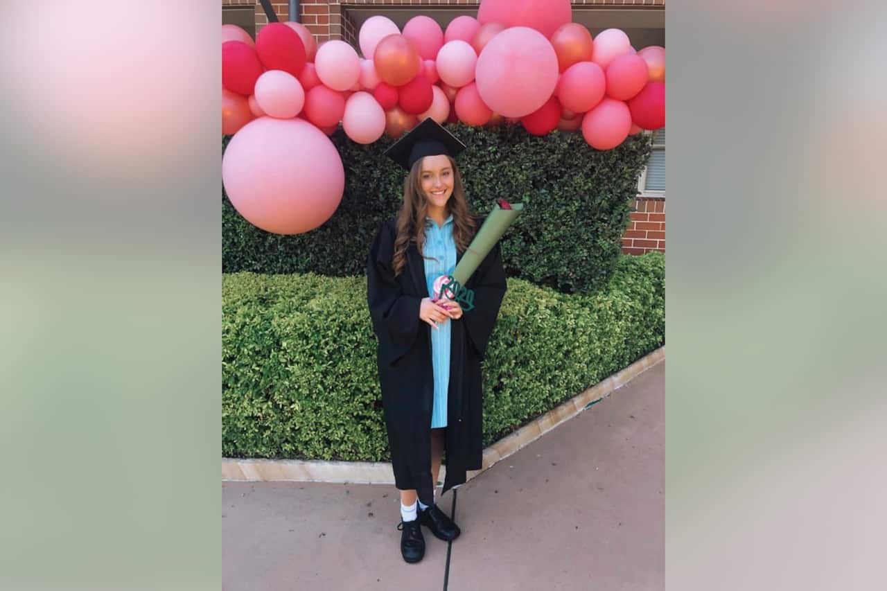 Young Caucasian woman with light brown hair posing for a photo in a black graduation cape and cap, with a light blue dress underneath. She is holding a small bunch of flowers and is standing in front of a green hedge decorated with pink balloons. 