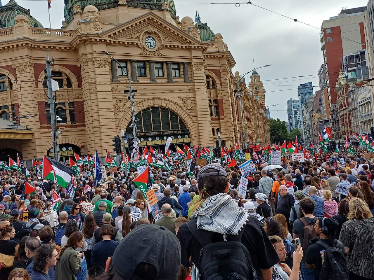 A large crowd of protesters carrying Palestinian flags and placards gathers in front of Flinders Street Station in Melbourne.