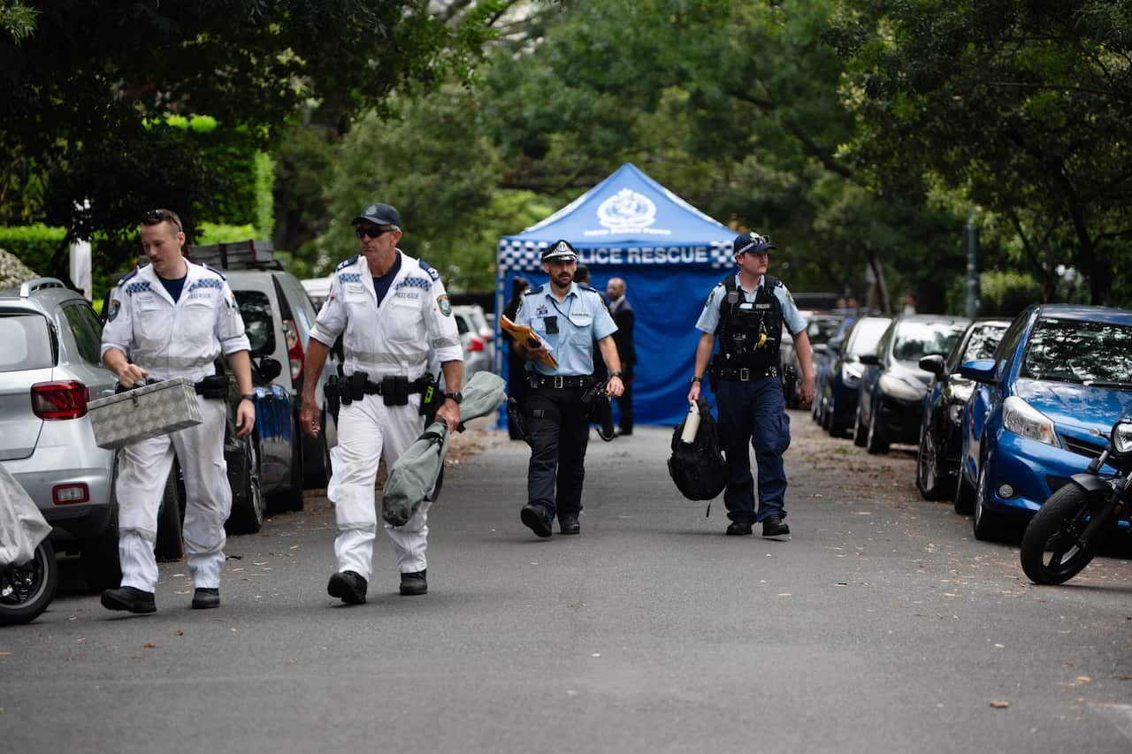 Four police officers, two wearing white forensic or rescue jumpsuits and two in standard blue uniforms, walk down a tree-lined residential street away from a blue "Police Rescue" tent.