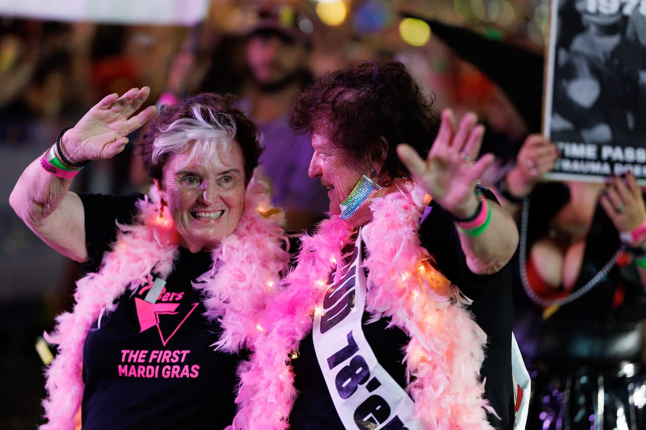 Two older women, known as "78ers," smile and wave during a night parade. They wear black T-shirts that say "The First Mardi Gras" and are draped in bright pink feather boas lit with fairy lights.