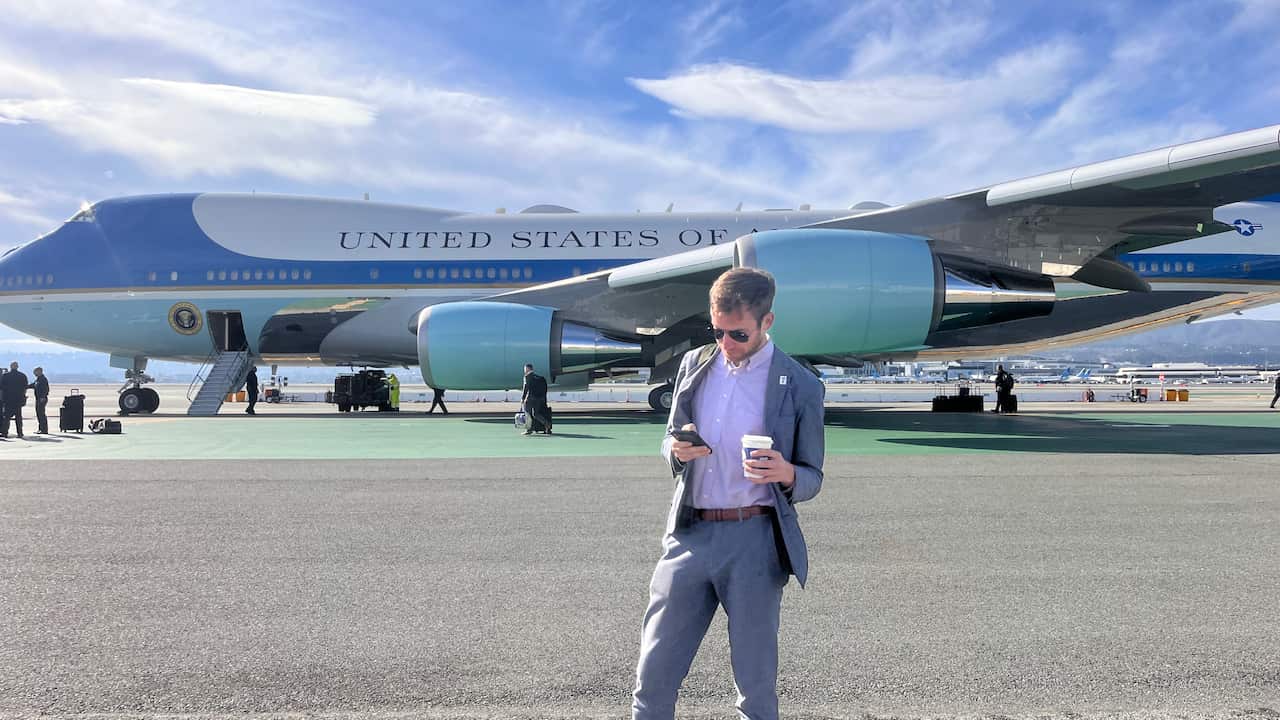 A man in a suit holding a coffee and looking at his phone. He is standing in front of a large blue and white aeroplane with United States of America written on its side