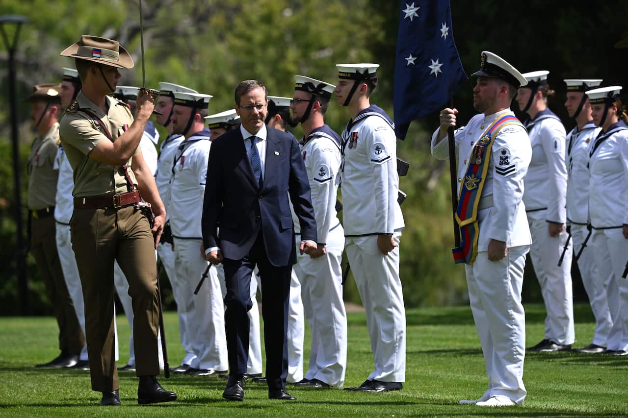 Isaac Herzog, wearing a dark suit, walks past a line of men in white uniforms. There is a man in khaki uniform and holding a sword up walking next to him