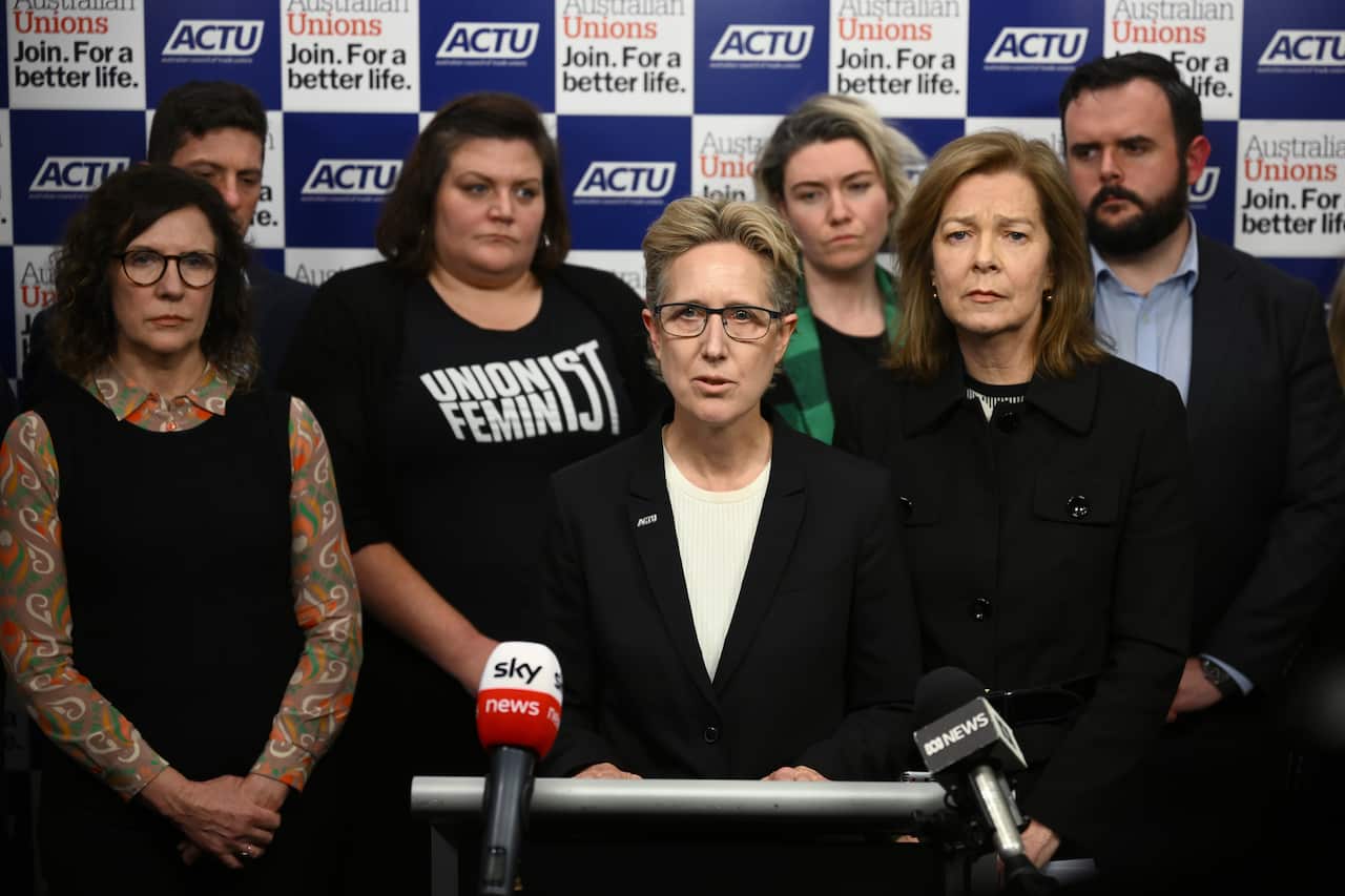 ACTU secretary Sally McManus standing at a lectern and speaking into microphones. Several other people are standing behind her.