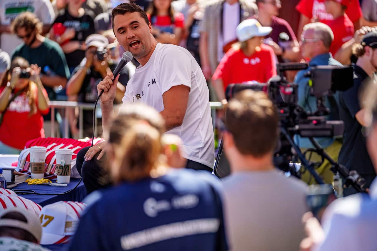 Charlie Kirk, sitting down and speaking into a microphone he's holding. There is a large crowd of people watching him.