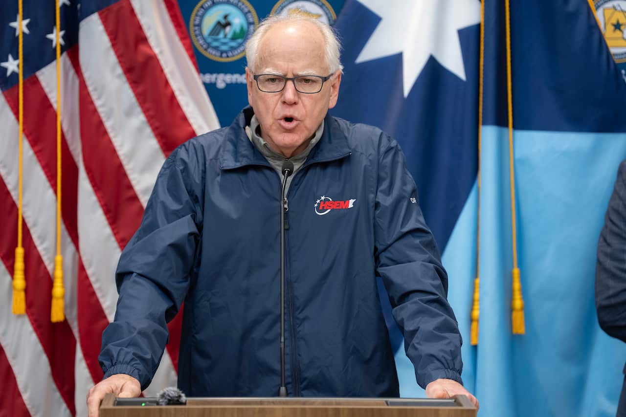 An older white man with balding white hair and glasses speaking at a lectern. An American flag hangs in the background. 