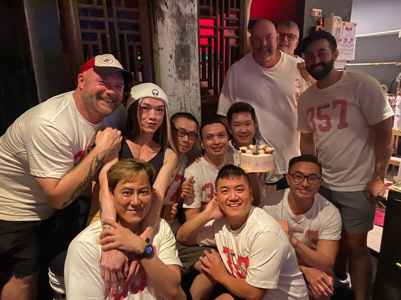 A photo of a group of queer and gay men huddling around a birthday cake inside a gay bar.
