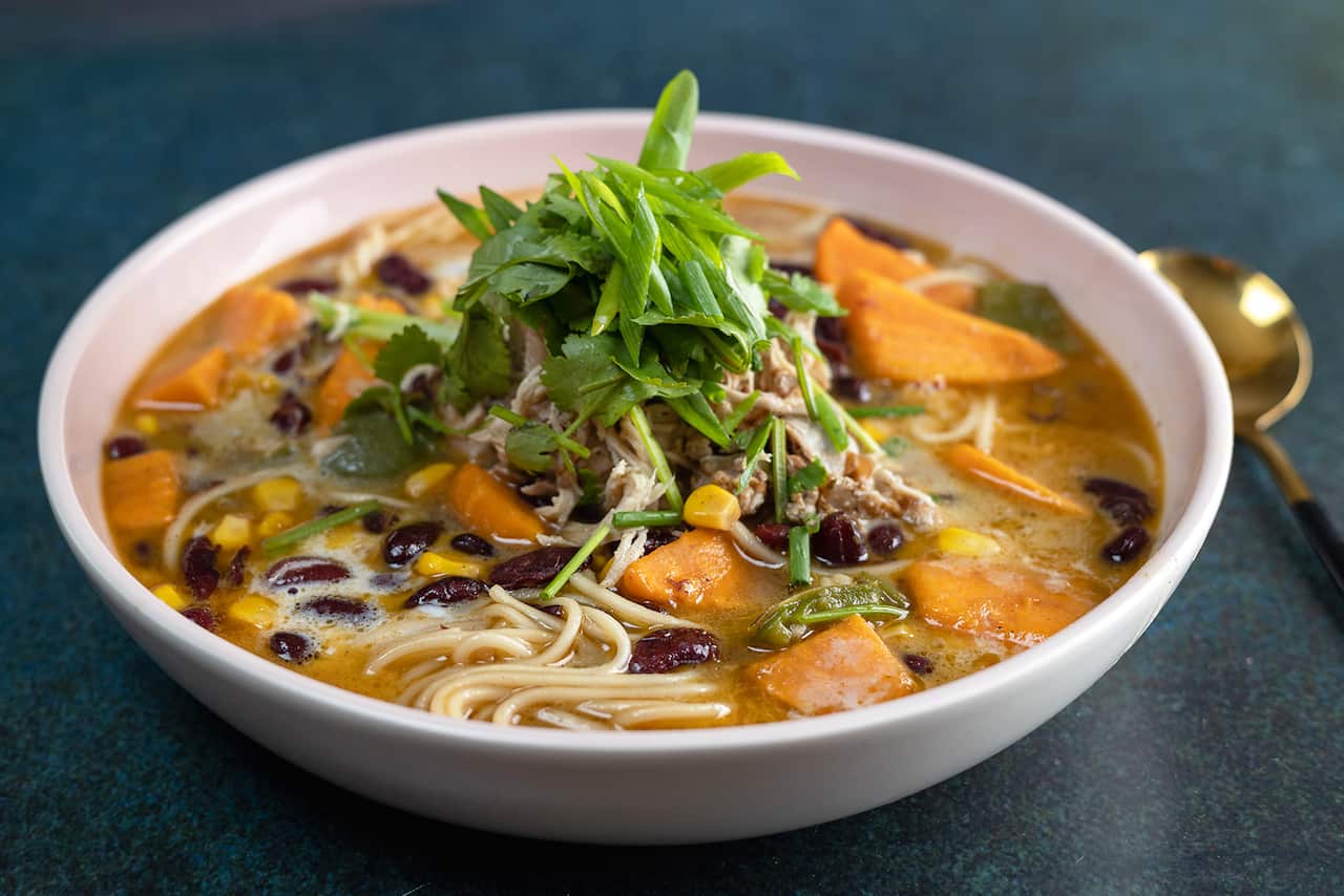 A white bowl holds noodle soup studded with sweet potato and topped with a pile of fresh coriander.