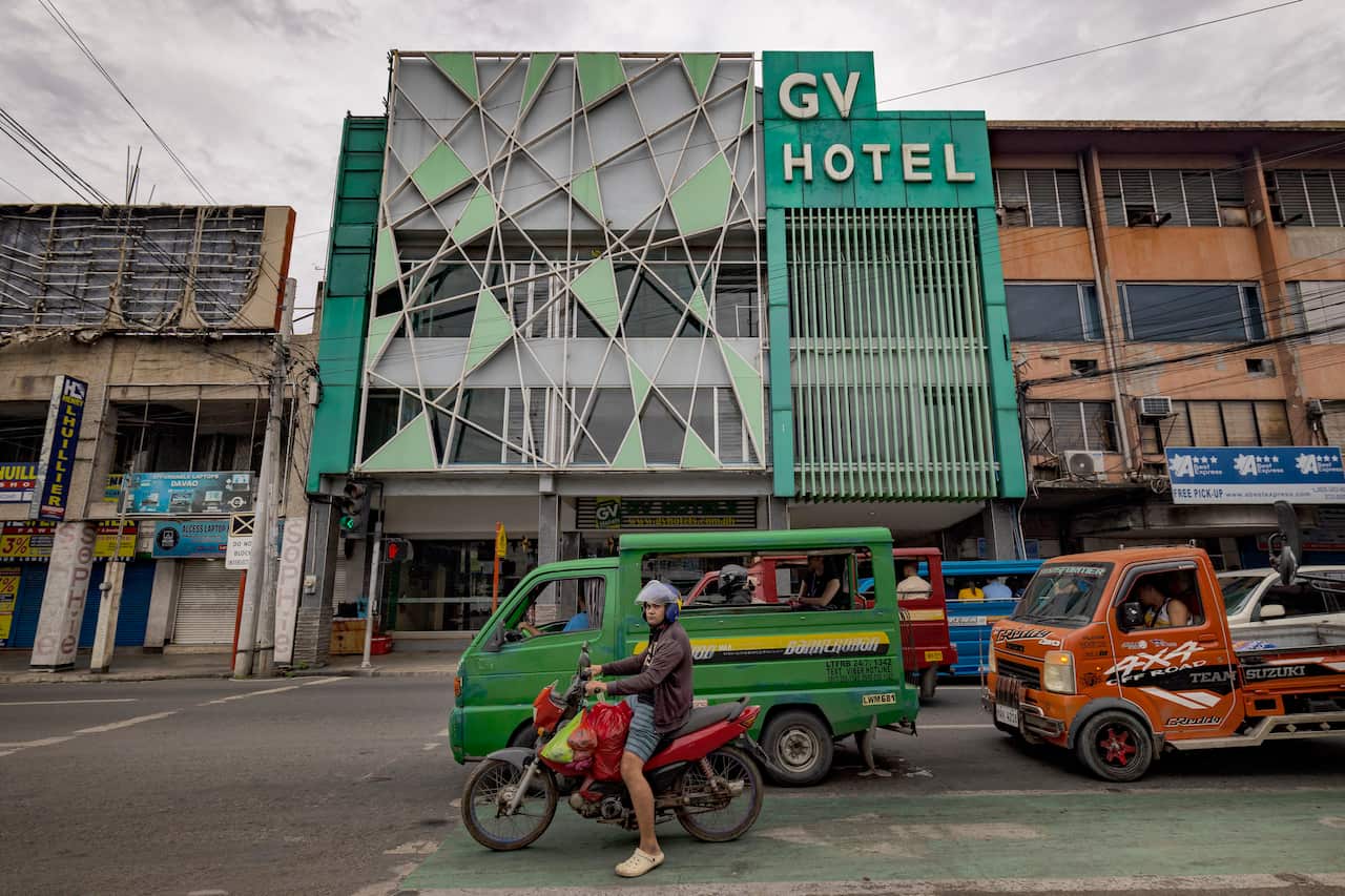 Pequenos caminhões em uma rua em frente a um pequeno prédio com fachada verde e branca e placa do GV ​​Hotel