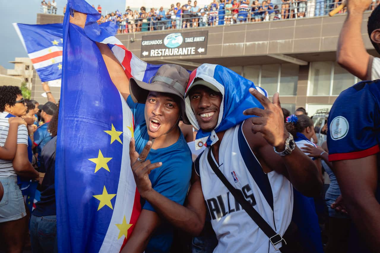 Two football fans celebrate their team's victory in a stadium. One of them is holding the Cape Verde flag while the other one has it tied like a scarf around his head.
