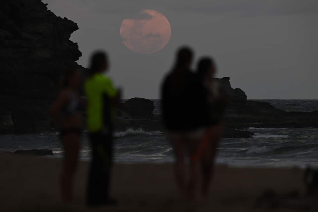 People on a beach are watching a large red moon rise over the ocean.